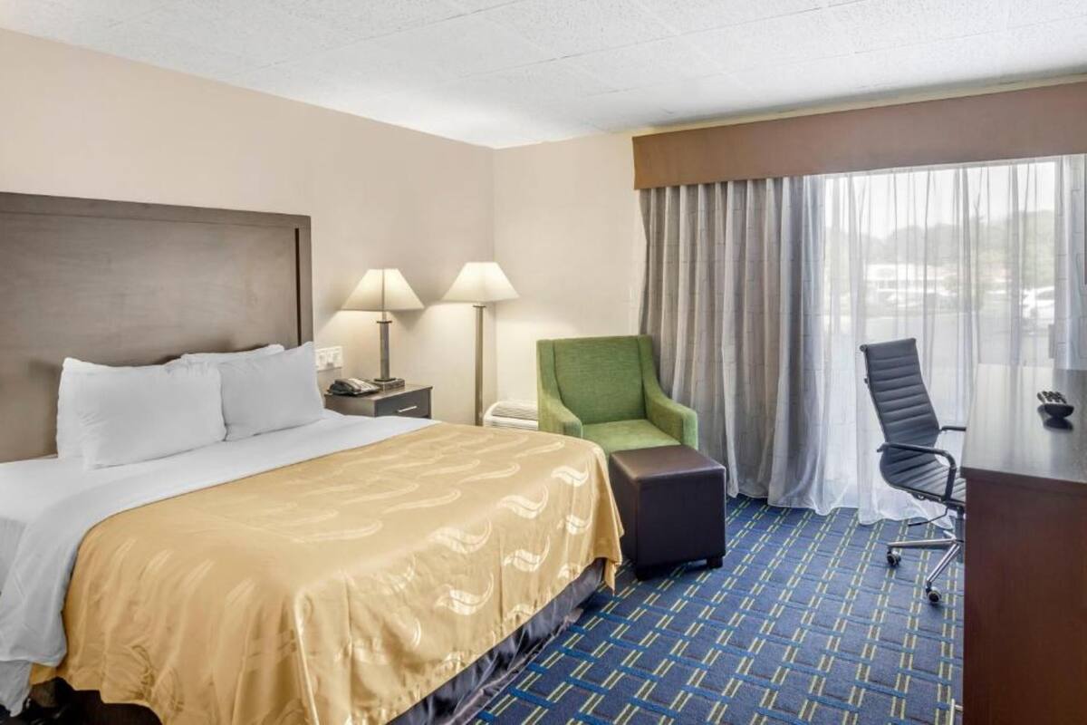 Una habitación de hotel con cama, dos lámparas, un sillón verde, una silla de oficina negra y un ventanal con cortinas.