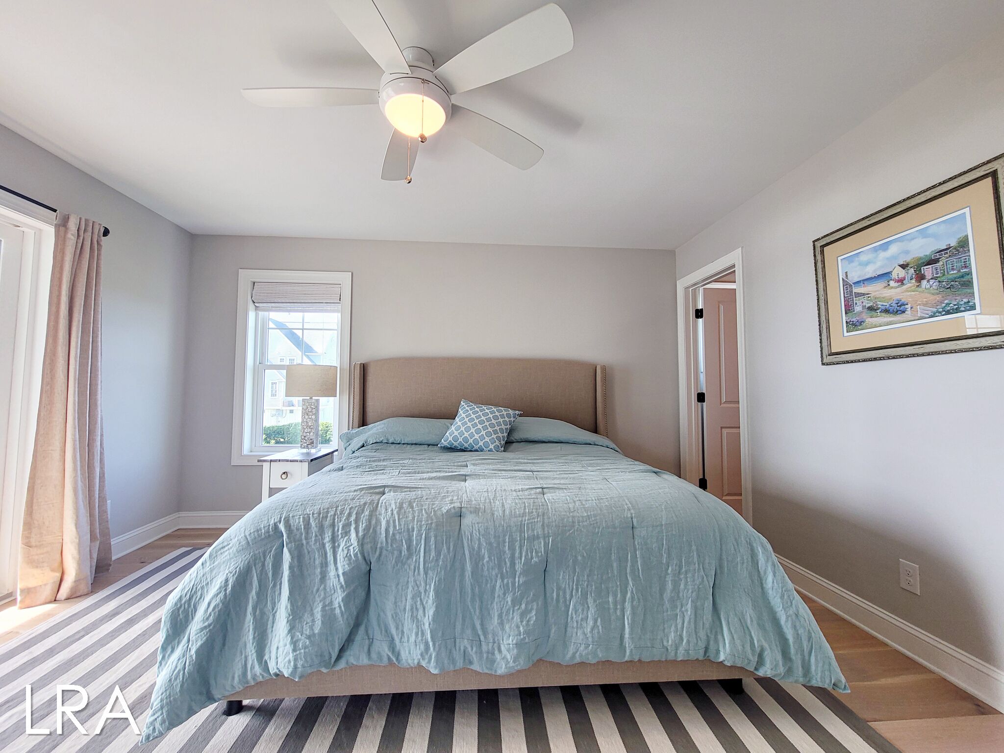 Ein Schlafzimmer mit einem Bett, einem Deckenventilator, einem Fenster mit Vorhängen, einem gerahmten Bild an der Wand und einem Streifenmuster-Teppich.