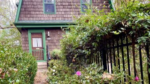 Une maison en bois avec une porte et des fenêtres vertes, entourée d’un jardin avec un chemin de gravier et une clôture noire.