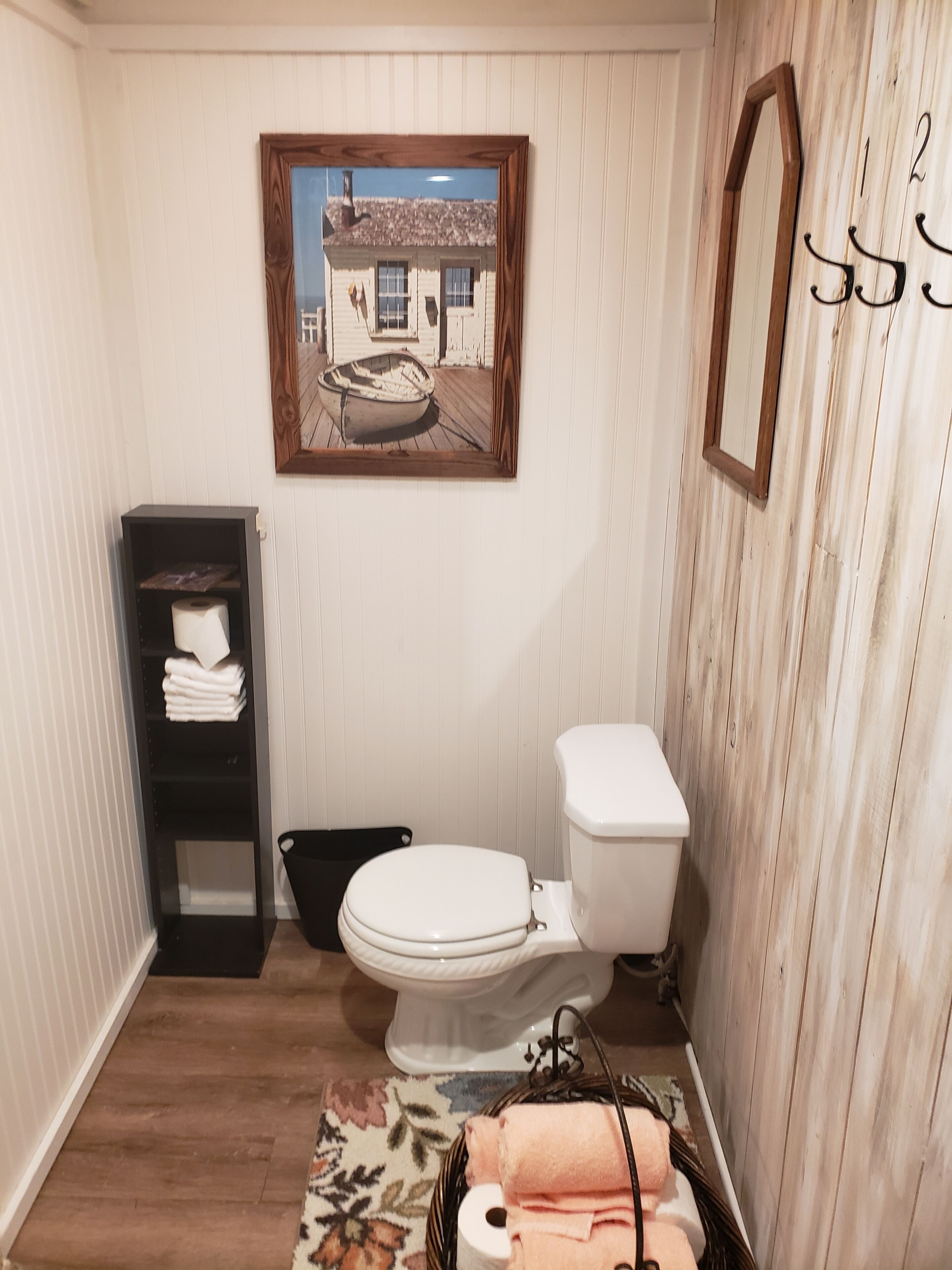 A bathroom with a toilet, a wooden floor, a shelf with towels, and a framed picture on the wall.