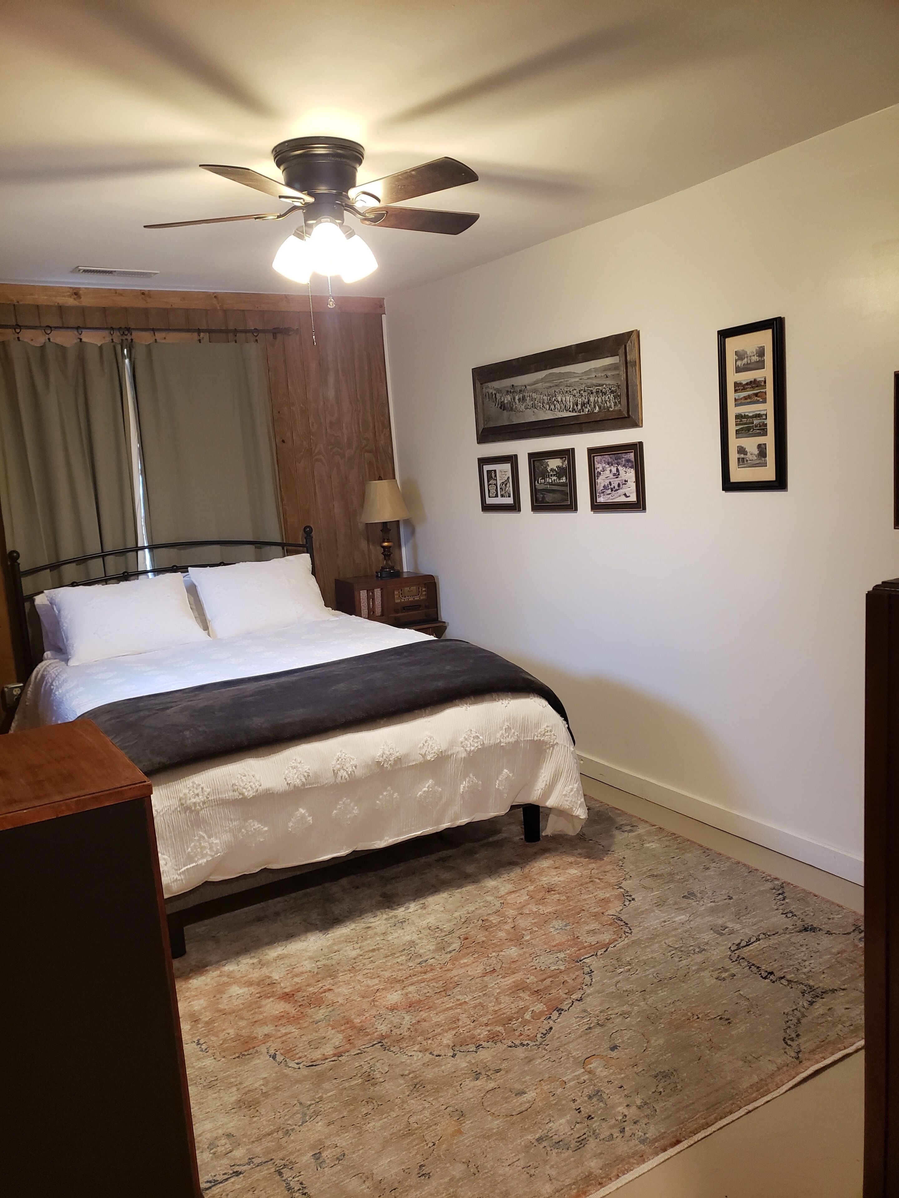 A bedroom with a bed, a ceiling fan, a nightstand, a lamp, and framed pictures on the wall.