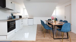 Een moderne keuken met witte kasten, een zwarte kookplaat en een eethoek met een houten tafel en blauwe stoelen.