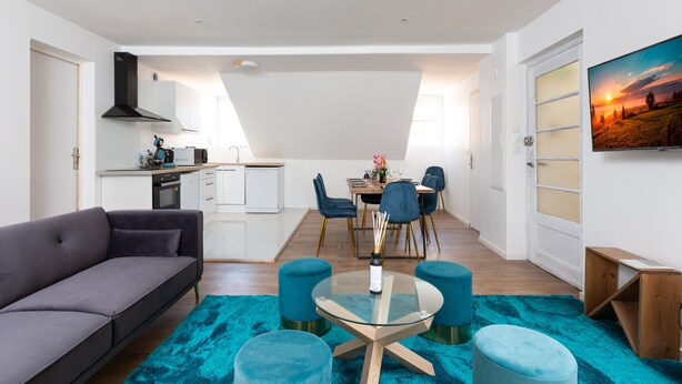 Een moderne woonkamer met een grijze bank, een glazen salontafel en turquoise voetenbankjes. Er is een eethoek met een tafel en stoelen, en een keuken op de achtergrond.