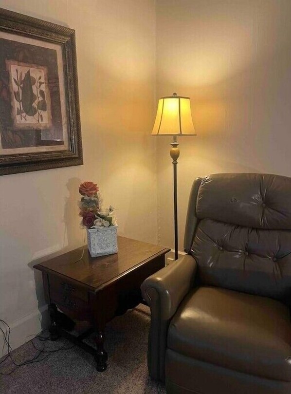 A room with a brown leather chair, a small wooden side table with a vase of flowers, a floor lamp, and a framed picture on the wall. - The Apartment (Weirton)