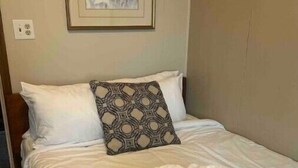 A neatly made bed with white linens, a patterned throw pillow, and a woven basket of folded towels. - The Apartment (Weirton)