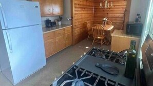 A compact kitchen with a gas stove, wooden cabinets, a refrigerator, and a dining area with chairs. - The Apartment (Weirton)