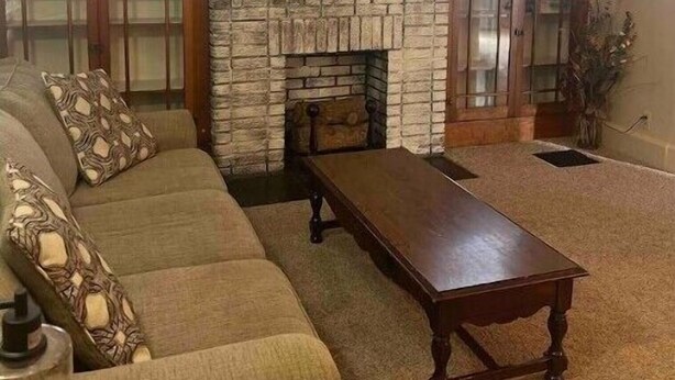A living room with a stone fireplace, a flat-screen TV mounted above it, a sofa with patterned cushions, and a wooden coffee table. - The Apartment (Weirton)