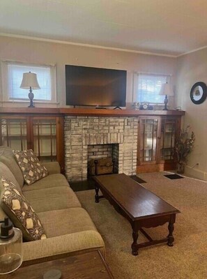 A living room with a stone fireplace, a flat-screen TV mounted above it, a sofa with patterned cushions, and a wooden coffee table. - The Apartment (Weirton)