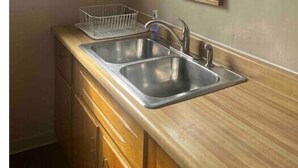 A kitchen with two stainless steel sinks, wooden cabinets, a window, and two framed artworks on the wall. - The Apartment (Weirton)