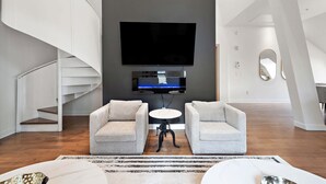 Un salon moderne avec un téléviseur à écran plat fixé à un mur foncé, deux fauteuils gris, une petite table ronde et un escalier en colimaçon sur la gauche.