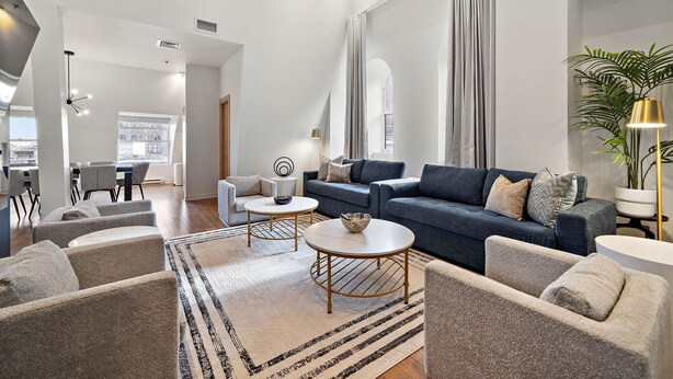 Un salon spacieux avec un canapé bleu foncé, des fauteuils beige et une table basse. On y trouve une grande fenêtre avec des rideaux, une plante en pot et une lampe.