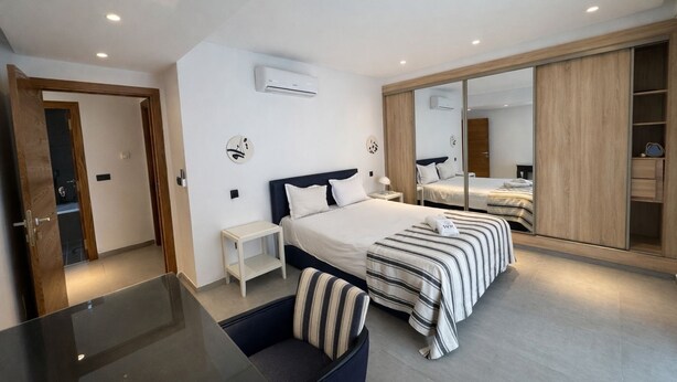 A modern bedroom with a large bed, a bedside table, a chair, a wardrobe, and an air conditioning unit. - Victor Hugo (La Marsa)