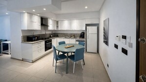 A modern kitchen with white cabinets, a granite countertop, a built-in oven, and a refrigerator. There is a dining area with a round table and blue chairs. - Victor Hugo (La Marsa)