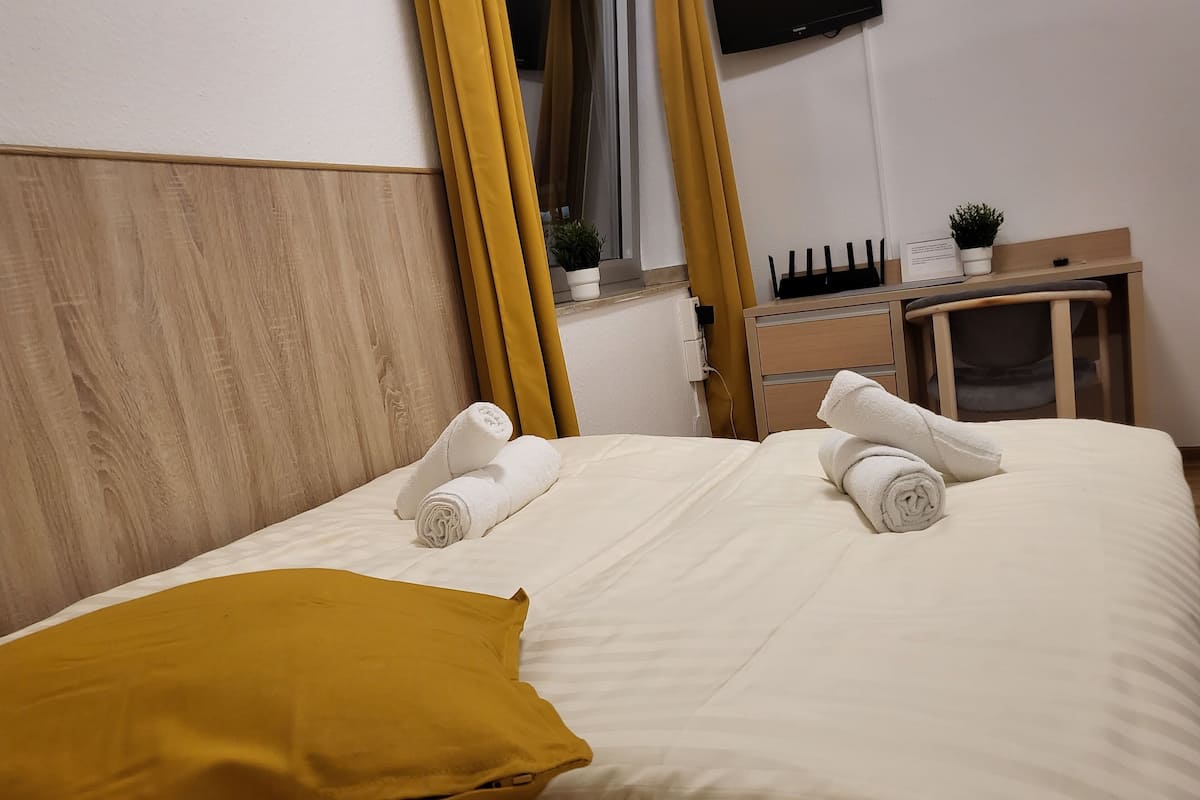 A neatly made bed with white linens and two rolled towels, a mustard-colored pillow, a wooden headboard, a bedside table with a plant, and a TV mounted on the wall.