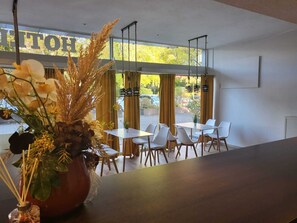 Un comedor moderno con sillas y mesas blancas, un jarrón grande con flores y vistas al exterior a través de amplias ventanas.