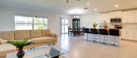 A modern living room with a glass coffee table, a beige sofa, and a dining area with a chandelier.