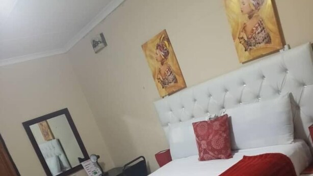Ein Schlafzimmer mit einem großen Bett, einem weißen Kopfteil und zwei roten Kissen. Ein Nachttisch mit Spiegel und ein kleiner Tisch mit einem roten Gegenstand darauf.