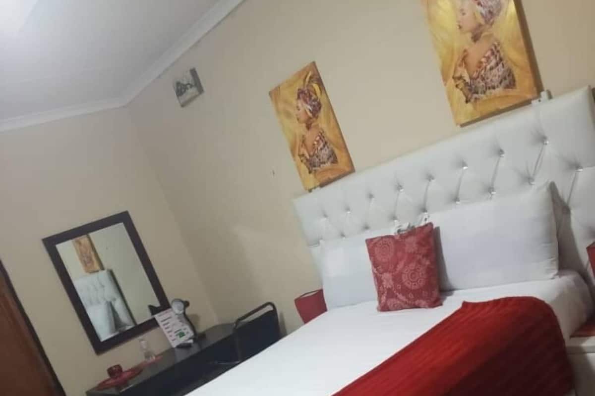 Un dormitorio con una cama grande, un cabecero blanco y dos almohadas rojas. Hay una mesita de noche con un espejo y una pequeña mesa con un objeto rojo encima.