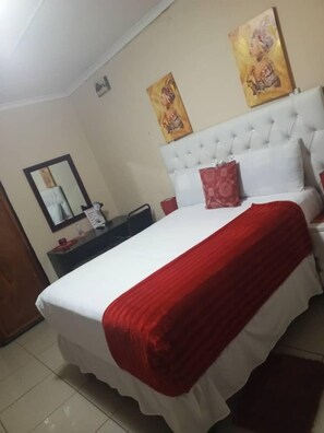 A bedroom with a large bed, a white headboard, and two red pillows. There is a nightstand with a mirror and a small table with a red object on it. - Beautiful gate guest house (Palapye)