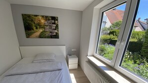 Une chambre à coucher comprenant un lit, une table de chevet, un radiateur et une fenêtre donnant sur un paysage verdoyant et des bâtiments.
