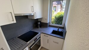 Une cuisine compacte dotée d’une cuisinière, d’un four et d’un évier intégrés, de meubles blancs et d’une fenêtre donnant sur un espace verdoyant.