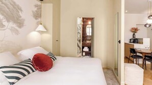 A bedroom with a bed, a red pillow, striped pillows, a hanging light, and a view into a dining area with a table and chairs.