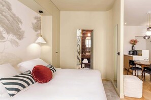 Quarto com cama, travesseiro vermelho, travesseiros listrados, luminária pendente e vista para uma sala de jantar com mesa e cadeiras.