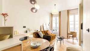 A modern living room with a wooden dining table, a sofa, and a small coffee table. There are two pendant lights, a vase with flowers, and a window with curtains.