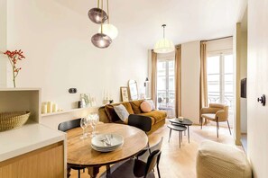Uma sala de estar moderna com uma mesa de jantar de madeira, um sofá e uma pequena mesa de centro. Há duas luminárias pendentes, um vaso com flores e uma janela com cortinas.