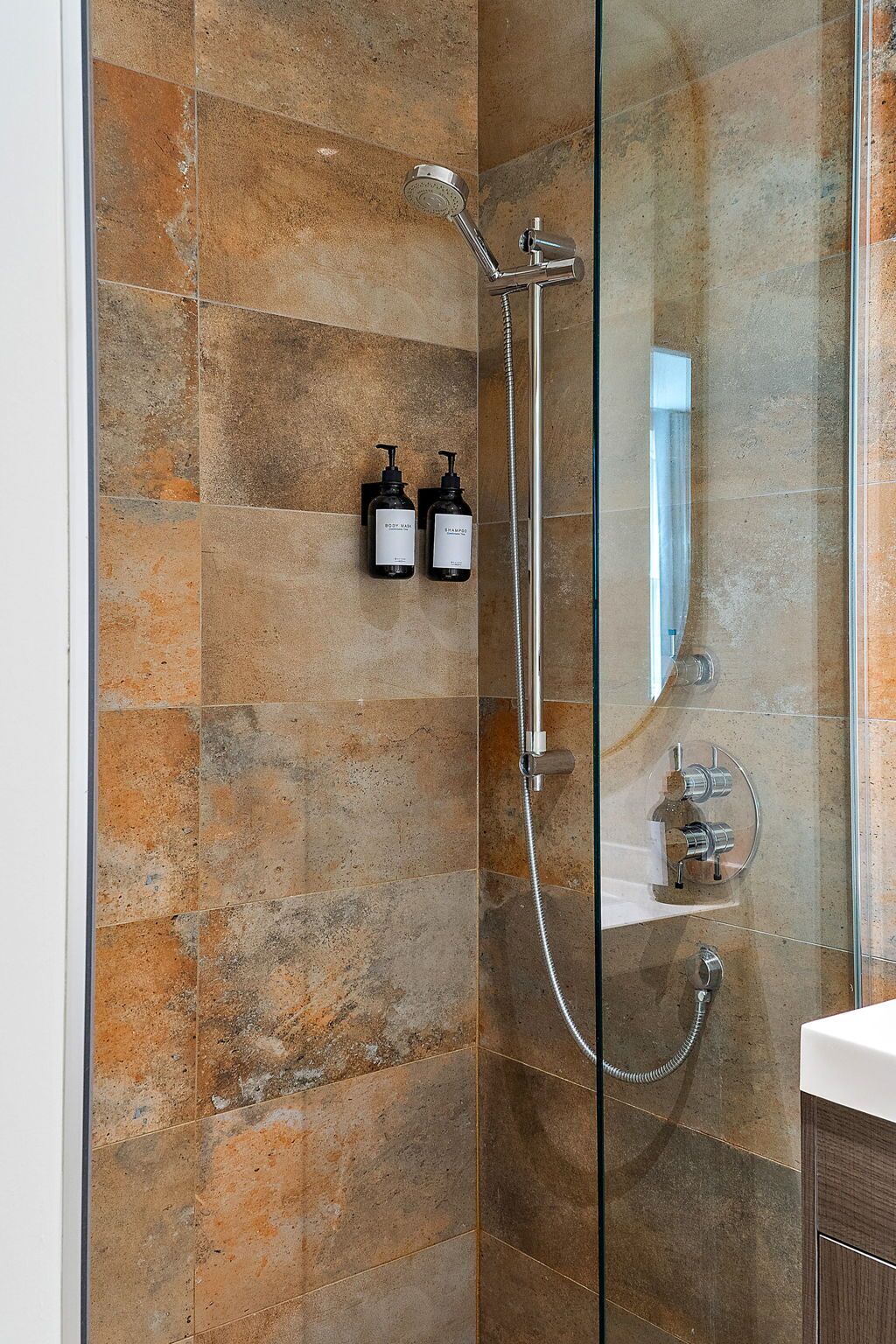 Eine moderne Dusche mit Glastrennwand, Handbrause und am Wand befestigtem Wasserhahn. Zwei Shampooflaschen sind an der Wand angebracht.