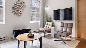 A modern living room with a round wooden table, two plush armchairs, a large flat-screen TV, and a decorative wall piece. - Nomade MTL, Old MTL, Luxury 2BD 2BA, St Jacques (Montréal)