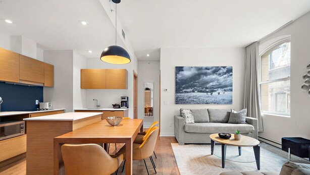 A modern kitchen with wooden cabinets, a dining area with a table and chairs, a living room with a sofa and a coffee table, and a large window with curtains. - Nomade MTL, Old MTL, Luxury 2BD 2BA, St Jacques (Montréal)