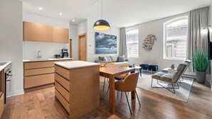 A modern kitchen with wooden cabinets, a dining area with a wooden table and chairs, and a living room with a sofa and a large window. - Nomade MTL, Old MTL, Luxury 2BD 2BA, St Jacques (Montréal)