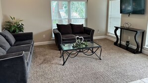 A living room with a dark-colored sofa, a glass coffee table, and a television mounted on the wall. - Unit B (Moss Point)
