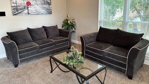A living room with two dark sofas, a glass coffee table with plants, a painting of a street scene, and a window with blinds. - Unit B (Moss Point)
