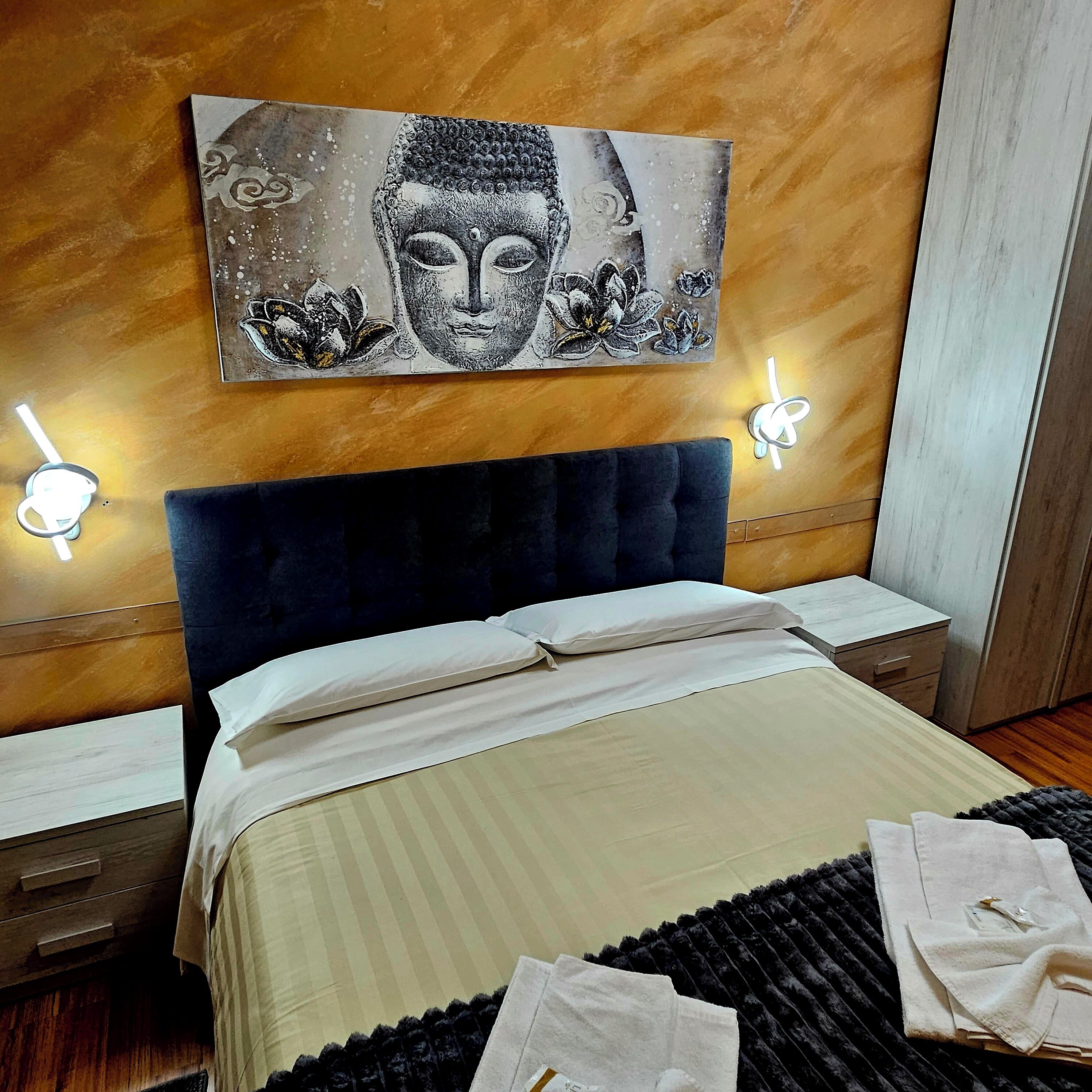 A bedroom with a large bed, a dark headboard, and a wall-mounted Buddha painting. There are two bedside tables with lamps.