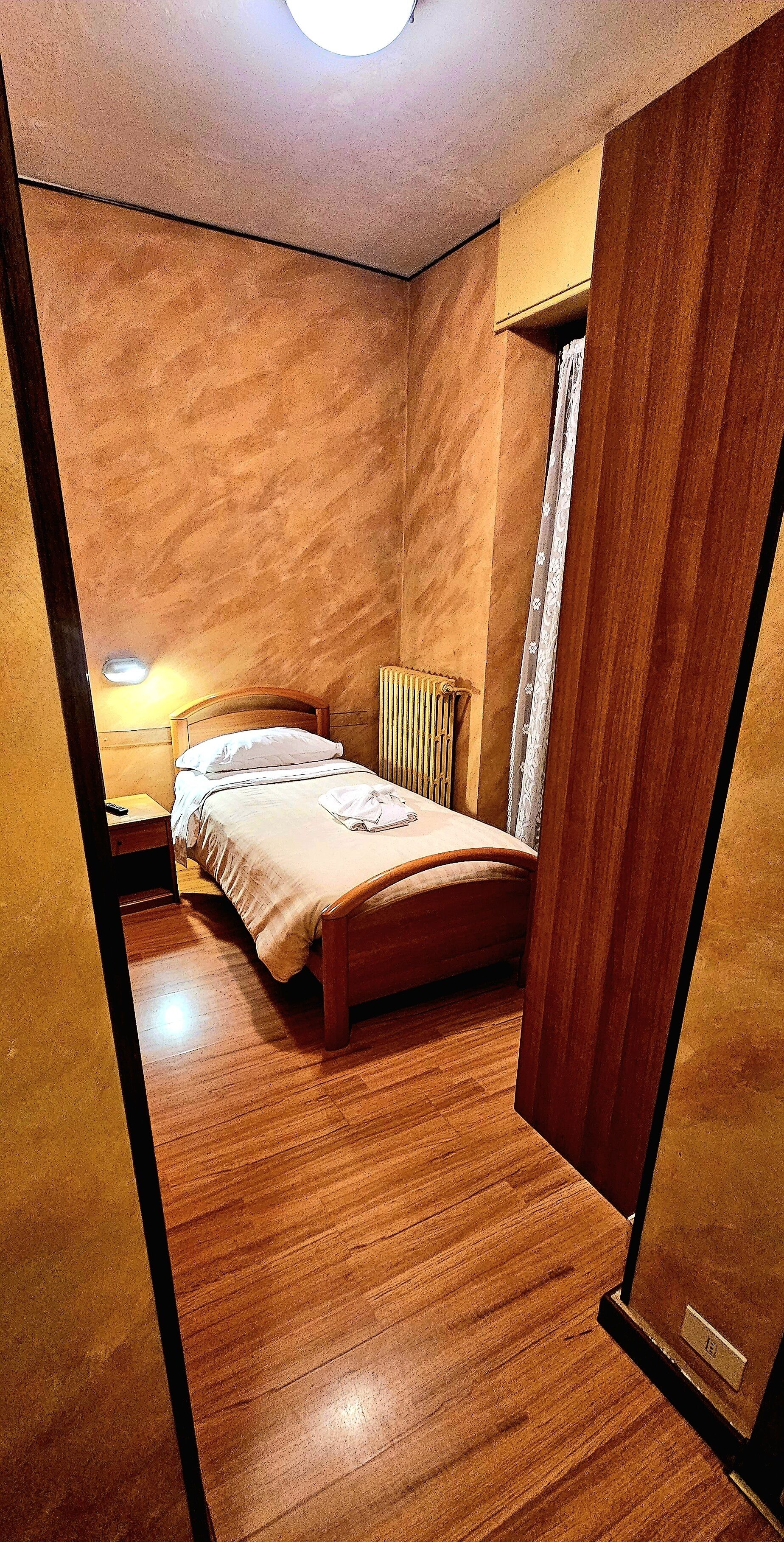 Ein Schlafzimmer mit Holzboden, einem Bett mit Kopfteil, einem Nachttisch, einer Lampe und einem Fenster mit Vorhängen.