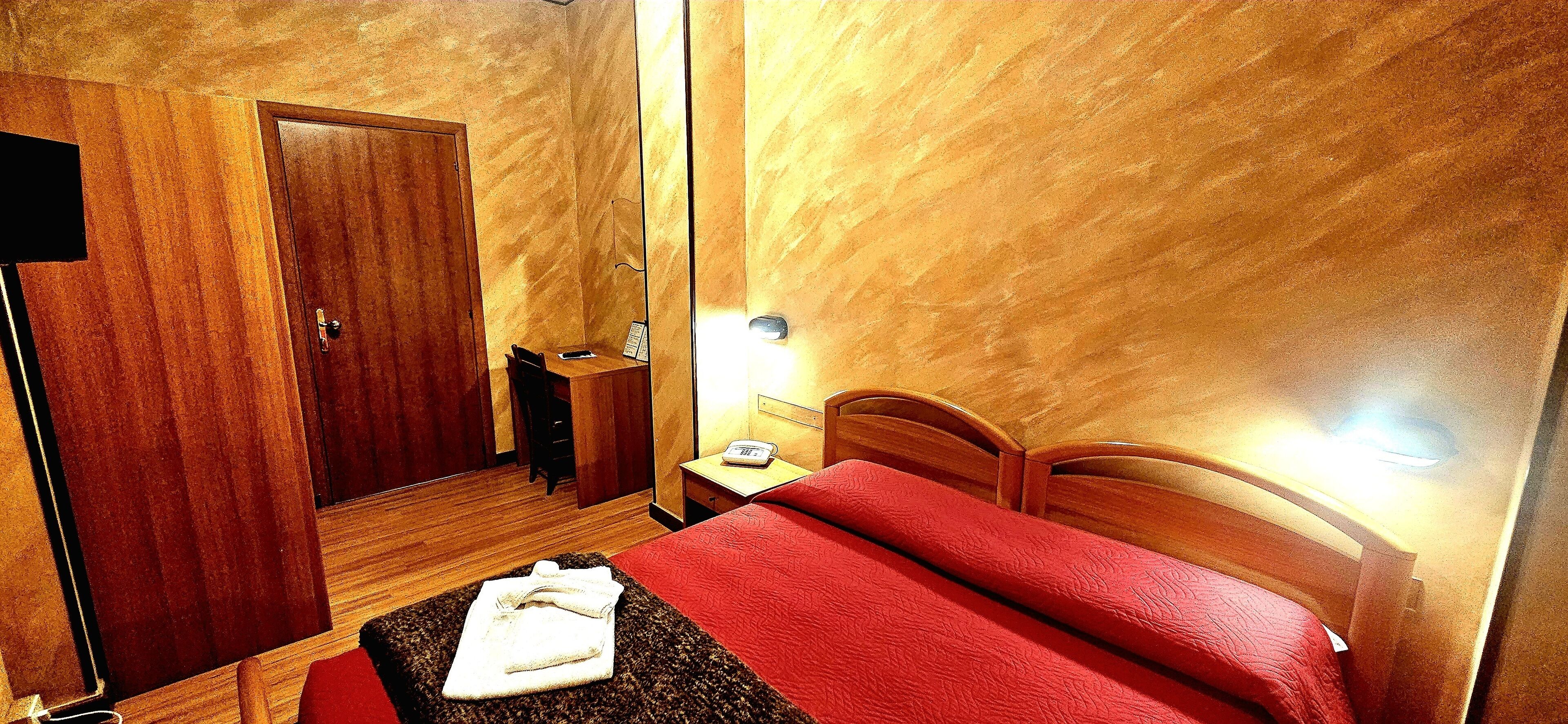 Una camera d'albergo con un armadio in legno, un comodino con una lampada, una coperta rossa e un asciugamano piegato sul pavimento.