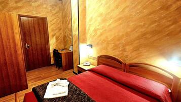 Una camera d'albergo con un armadio in legno, un comodino con una lampada, una coperta rossa e un asciugamano piegato sul pavimento.