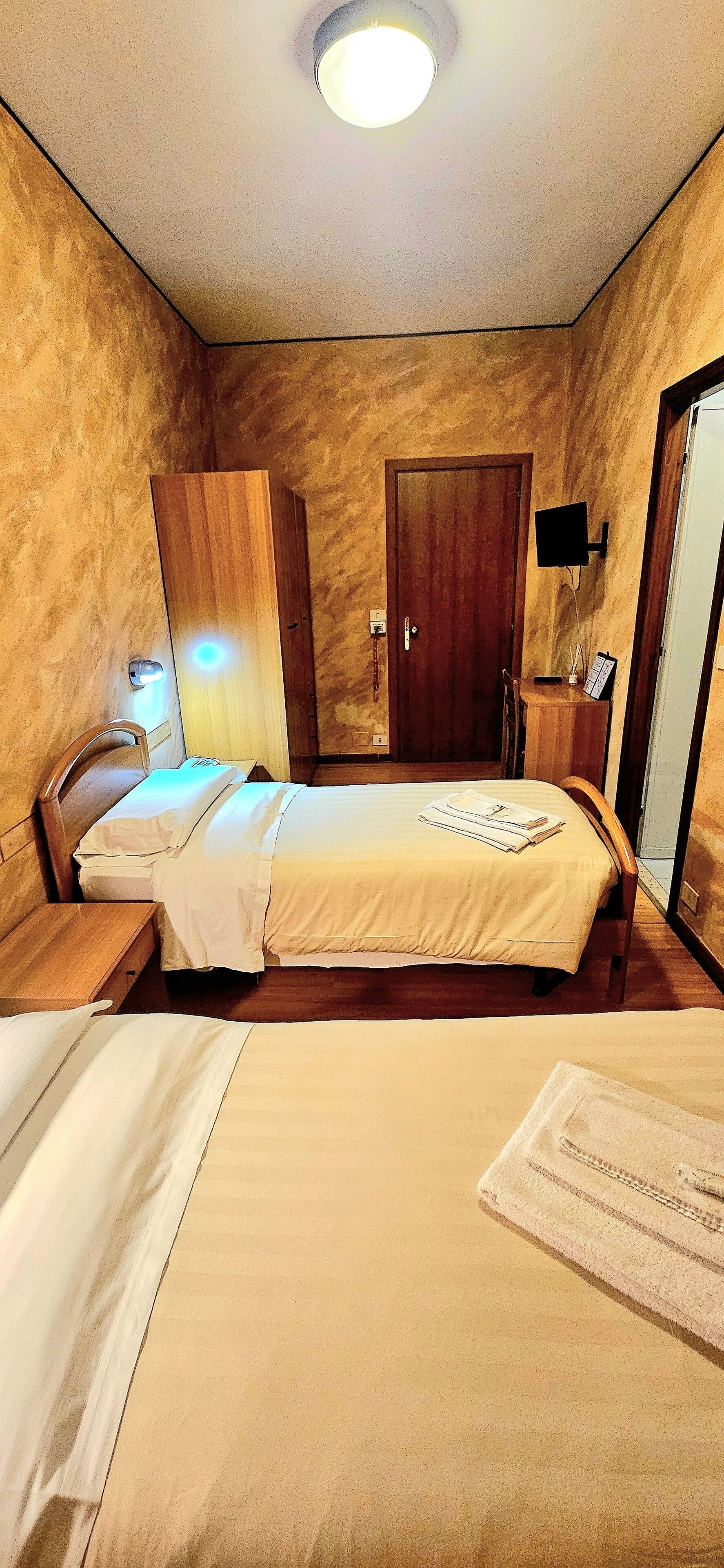 A hotel room with a bed, a nightstand, a wooden wardrobe, a mirror, and a TV mounted on the wall.