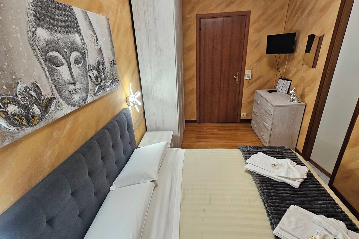 A bedroom with a tufted headboard, a large bed with white linens, a dark patterned rug, a wooden wardrobe, a wooden door, and a wall-mounted flat-screen TV.