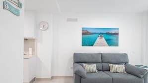A modern living room with a grey sofa, a clock, a framed picture of a pier, and a small shelf with a candle. - Casa Arrullo Las Canteras - next to the beach (Las Palmas de Gran Canaria)