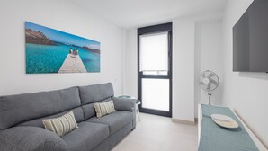 A modern living room with a grey sofa, a small side table, a fan, and a large window with blinds. - Casa Arrullo Las Canteras - next to the beach (Las Palmas de Gran Canaria)
