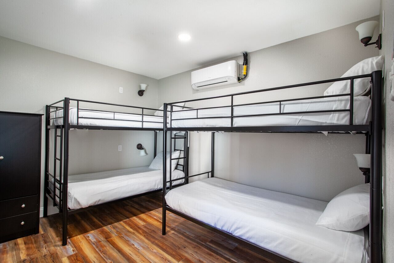 Une chambre de dortoir avec trois lits superposés, un plancher en bois, une armoire noire, un climatiseur fixé au mur et une lampe murale.