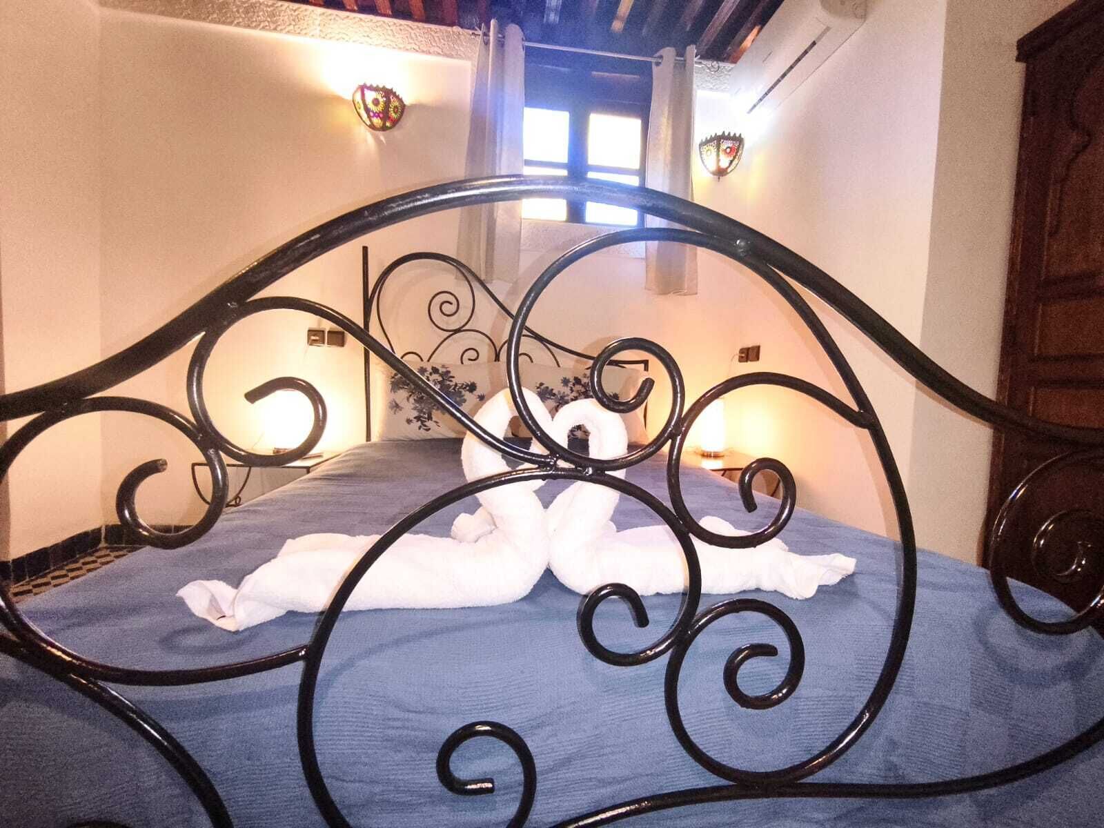 Uma cama com estrutura de metal preto e colcha azul, uma toalha branca e uma pequena mesa de cabeceira com abajur.