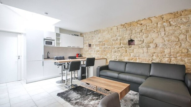 A modern living room with a stone wall, a black leather sofa, a wooden coffee table, and a kitchen area with white cabinets and a microwave. - 01.Duplex near Tour Eiffel & Montparnasse Station (Paris)