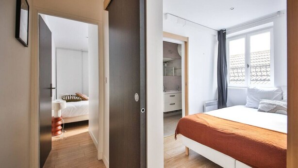 A bedroom with a bed, a window with curtains, a wardrobe, and a bathroom visible through an open door. - 02.Duplex#2Bedrooms#Sèvres-Lecourbe#Champ-de-Mars (Paris)