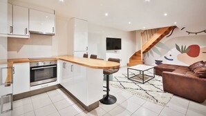 A modern kitchen with white cabinets, a wooden countertop, and an oven. There is a bar stool, a brown leather sofa, and a TV mounted on the wall. - 02.Duplex#2Bedrooms#Sèvres-Lecourbe#Champ-de-Mars (Paris)