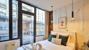 Moderni hotellihuone, jossa on suuri sänky, lattiasta kattoon ulottuvat ikkunat, joista avautuu näkymä kaupunkiin, seinällä oleva taideteos ja viihtyisä tunnelma.