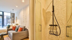 Moderni hotellihuone, jossa on sohva, lasinen suihkuseinä ja suuri ikkuna, josta avautuu näkymä kaupunkiin.